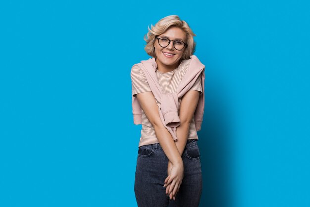 Blonde woman wearing glasses is feeling happy on a blue studio wall with glasses smiling