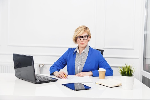 Blonde woman wears blue jacket manage with documents on the table with flowerpot white office room busy working concept