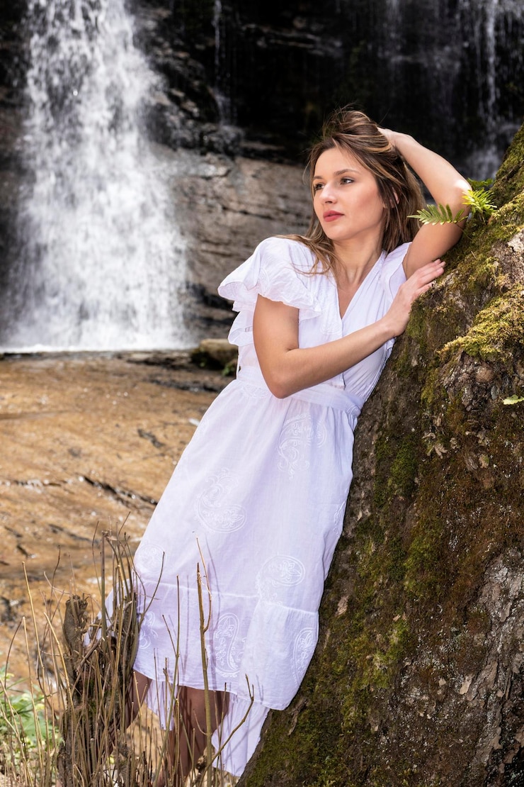 Photo blonde woman in a white dress leaning on a tree in a waterfall