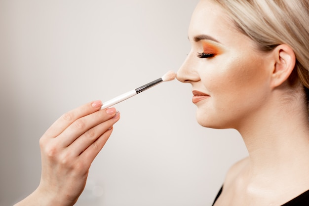 Blonde woman with bright makeup in profile. In the frame, the makeup artist's hand holding a makeup brush. Blonde model smiling