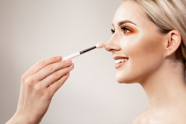 Blonde woman with bright makeup in profile. In the frame, the makeup artist's hand holding a makeup brush. Blonde model smiling