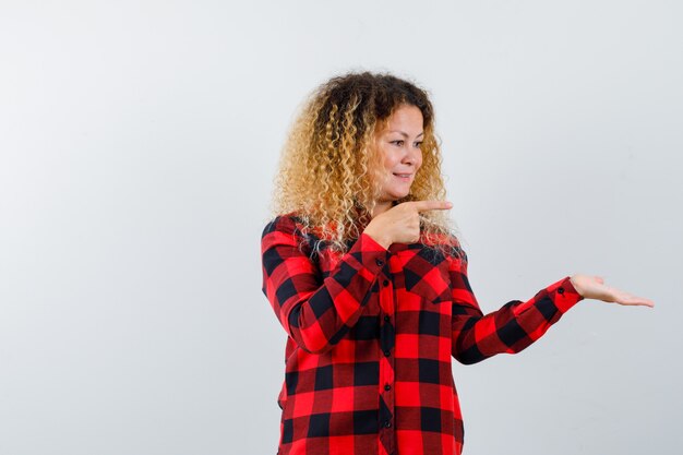 Blonde woman with curly hair in checked shirt pointing at her palm spread aside and looking cheery , front view.