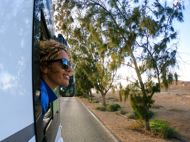 Blonde woman with curly hair looks out of the window of the moving camper Female tastes the wind in her face and smiles Travel and tourism concept