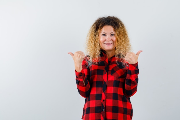 Blonde woman with curly hair pointing to the opposite directions with thumbs in checked shirt and looking cheery , front view.