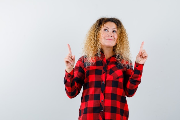 Blonde woman with curly hair pointing up in checked shirt and looking thankful , front view.