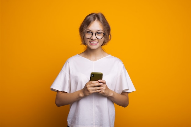 Donna bionda con gli occhiali in chat sul cellulare e sorridere su un muro giallo che indossa una maglietta bianca