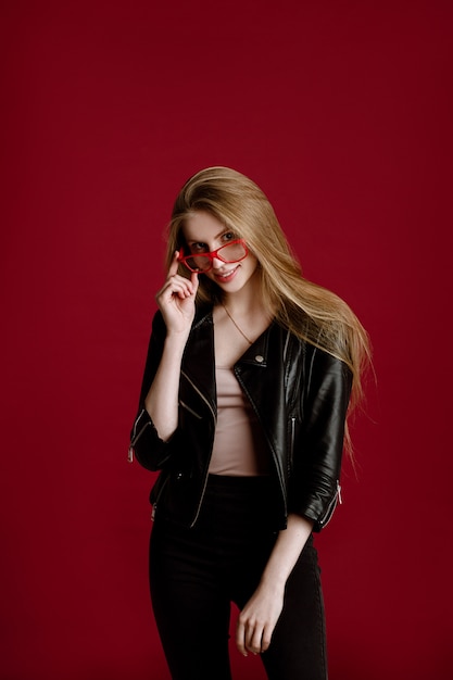 A blonde woman with long hair looks confidently at the camera with glasses on a red background