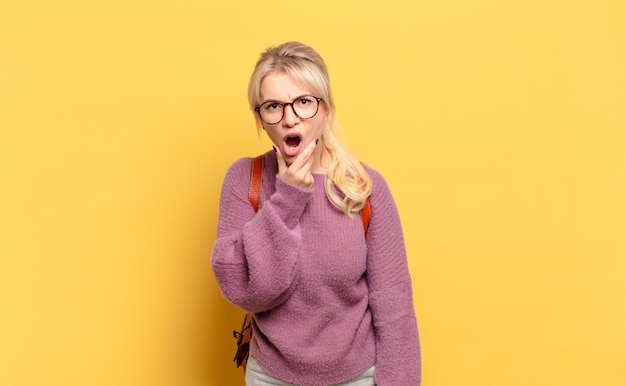 Blonde woman with mouth and eyes wide open and hand on chin, feeling unpleasantly shocked, saying what or wow