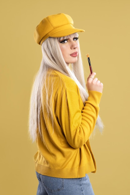 Blonde woman with painter's cap and brush looking towards camera, yellow background