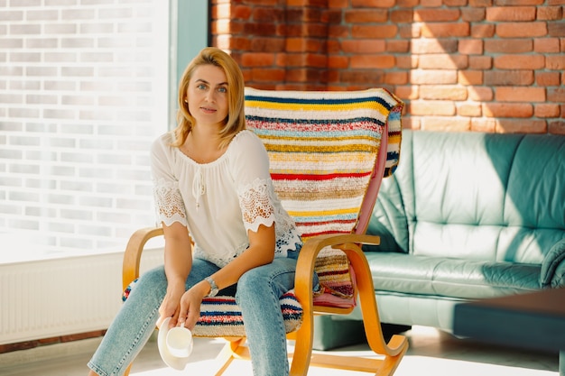 A blonde woman with a pleasant appearance sits in a rocking chair after drinking a cup of tea