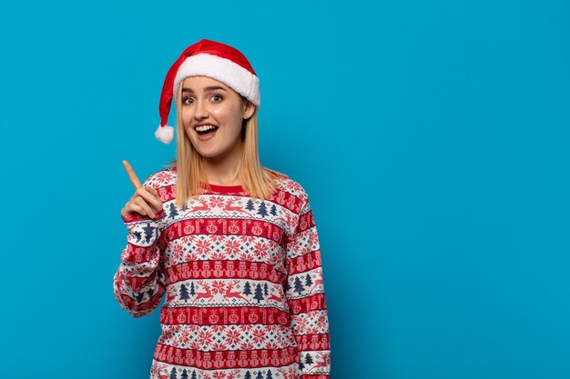 Blonde woman with santa hat feeling like a happy and excited genius after realizing an idea, cheerfully raising finger, eureka!