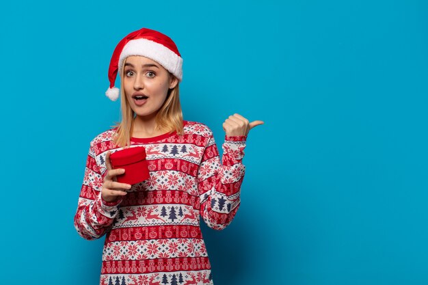 Blonde woman with santa hat looking astonished in disbelief, pointing at object on the side and saying wow, unbelievable