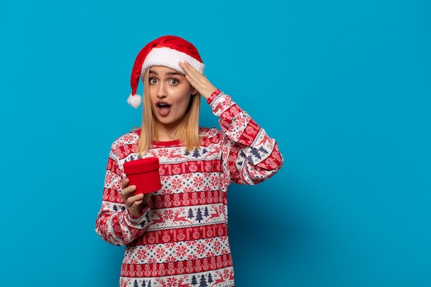 Donna bionda con cappello da babbo natale che sembra felice, stupita e sorpresa, sorridente e realizzando incredibili e incredibili buone notizie