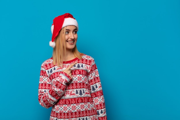 Blonde woman with santa hat smiling cheerfully, feeling happy and pointing to the side and upwards, showing object in copy space