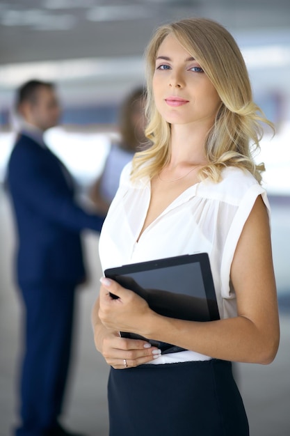 Blonde woman with touchpad computer looking at camera and smiling while business people shaking hands over background