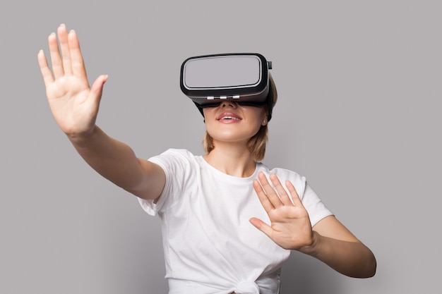 Blonde woman with vr headset is touching something and posing on a grey studio wall