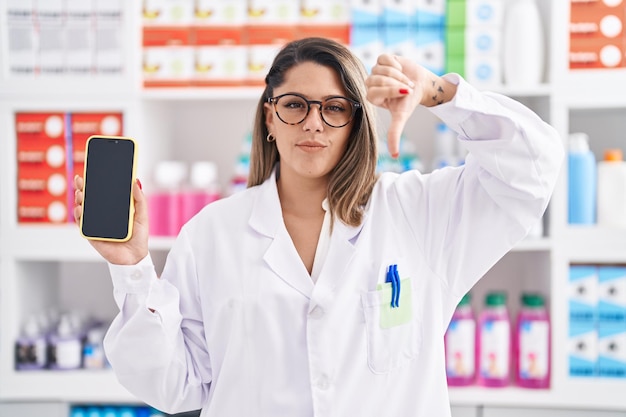 Blonde woman working at pharmacy drugstore showing smartphone screen with angry face negative sign showing dislike with thumbs down rejection concept