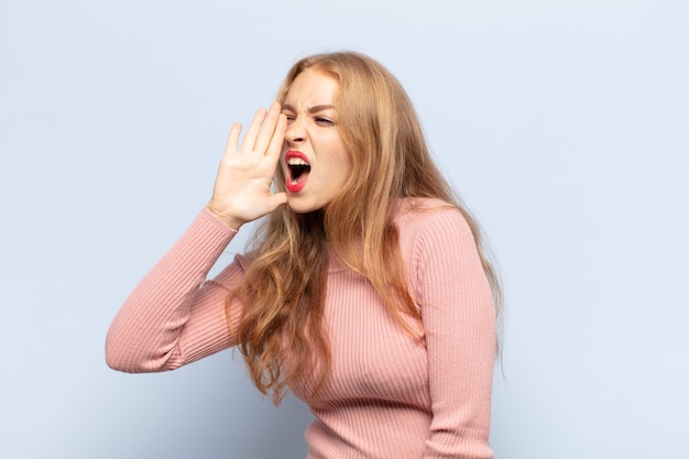 Blonde woman yelling loudly and angrily to copy space on the side, with hand next to mouth