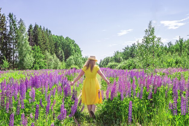Blonde woman in a yellow dress in lupine field. Summer sunny day.