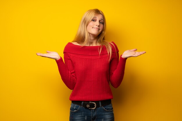 Blonde woman over yellow wall having doubts while raising hands and shoulders