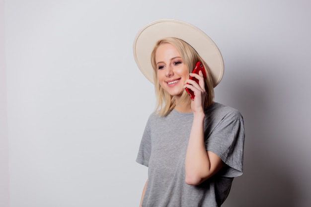 Blonde womanin hoed die mobiele telefoon op witte muur met behulp van