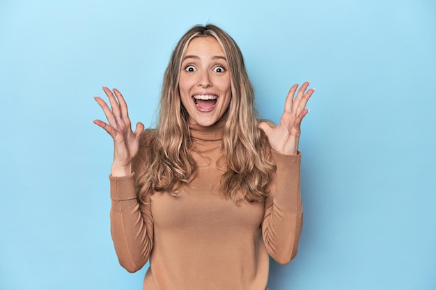 Blonde young caucasian woman in blue studio celebrating a victory or success