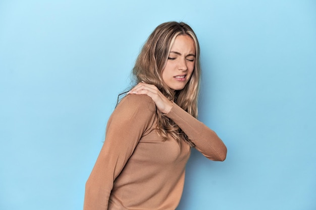 Blonde young caucasian woman in blue studio having a shoulder pain