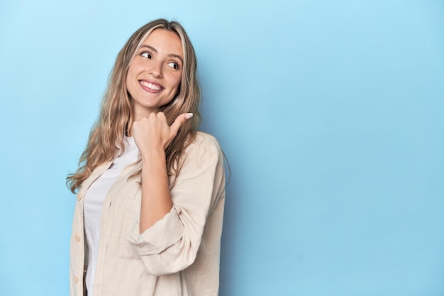 Blonde young caucasian woman in blue studio points with thumb finger away laughing and carefree