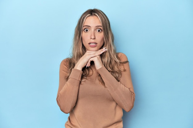 Blonde young caucasian woman in blue studio praying for luck amazed and opening mouth