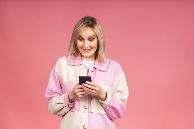 Blonde young female with terrible expression in smartphone, reads shocking news on web page, isolated on pink background. Shocked amazed woman using mobile phone.