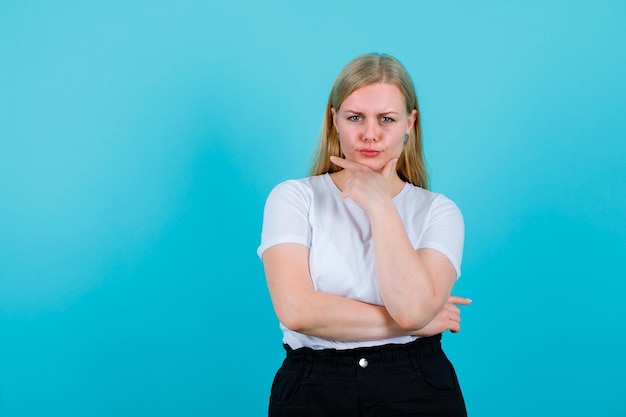 ブロンドの若い女の子は青い背景のあごの下で手を握ってbuを考えています