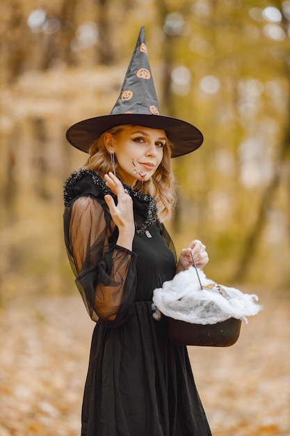 Blonde young girl witch in forest on halloween. girl wearing\
black dress and cone hat. witch holding a magician stuff.