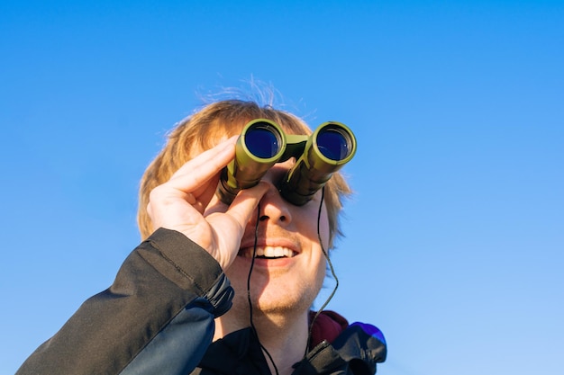 Blonde young man looking through binoculars while outdoors in nature Guy looking through binoculars on the blue sky Travel and wildlife concept