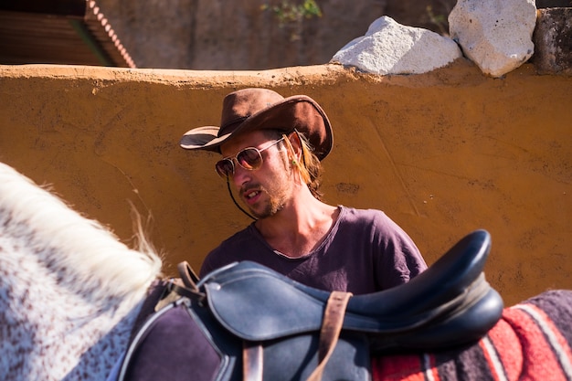 Blonde young man prepare a horse to go on excursion with cowboy hat.