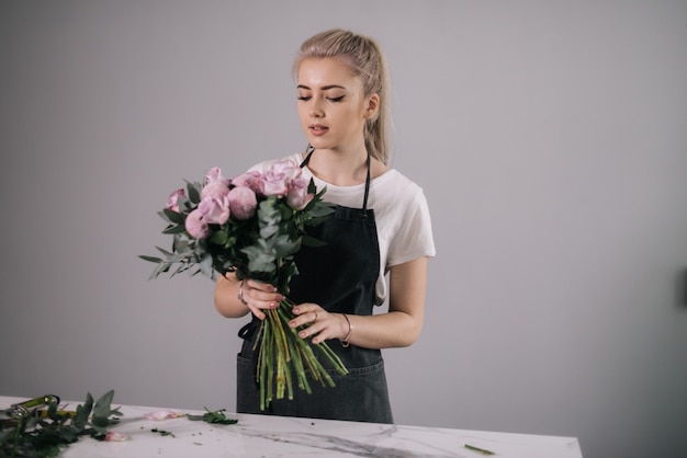 テーブルで花の花束と立っているエプロンを身に着けている金髪の若い女性の花屋