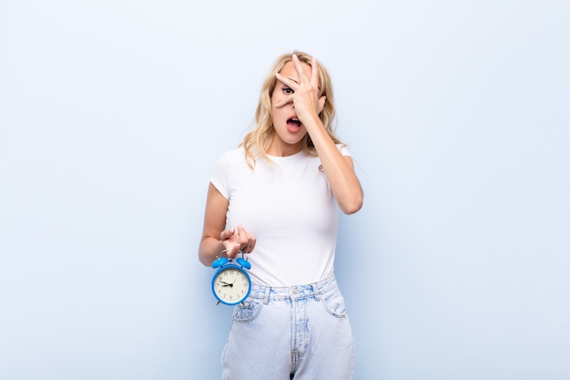 blonde young woman holding a clock looking shocked or terrified covering face with hand