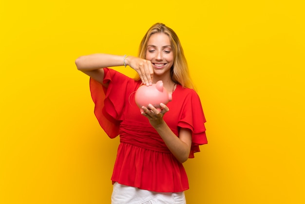 Blonde young woman over isolated yellow wall holding a big piggybank