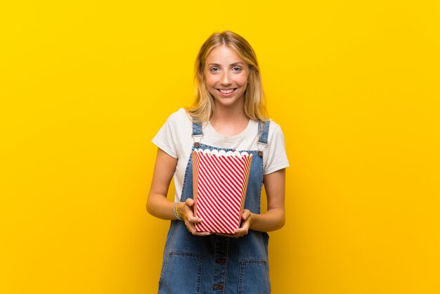 Blonde young woman over isolated yellow wall holding a bowl of popcorns