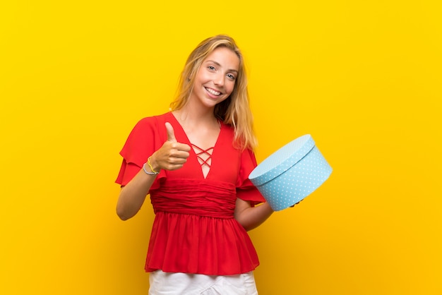 Blonde young woman over isolated yellow wall holding gift box