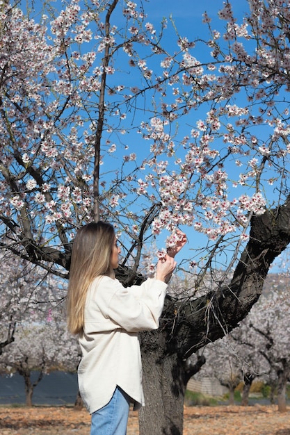 アーモンドの木の下で白とピンクの花を拾う金髪の若い女性