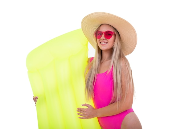 A blonde young woman in a pink swimsuit and hat holds air mattress