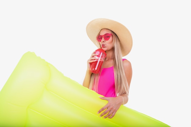 A blonde young woman in a pink swimsuit and hat holds air mattress
