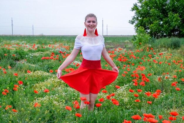 赤いスカートと白いシャツの赤いイヤリングを着た金髪の若い女性がケシ畑の真ん中にいる