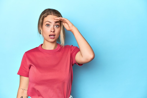 Photo blonde young woman in red tshirt on blue background shouts loud keeps eyes opened and hands tense