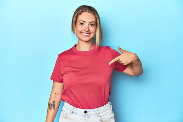 Blonde young woman in red tshirt person pointing by hand to a shirt copy space proud and confident