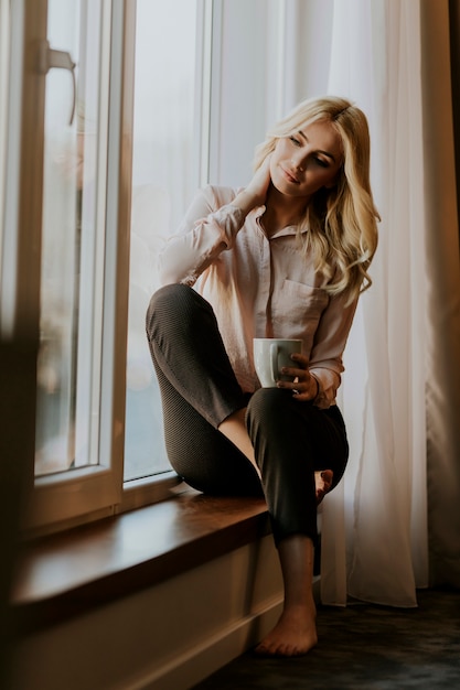 Blonde young woman relaxing and  drinking coffee by window