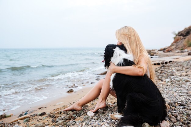 Giovane donna bionda che si siede e che abbraccia un cane sulla spiaggia