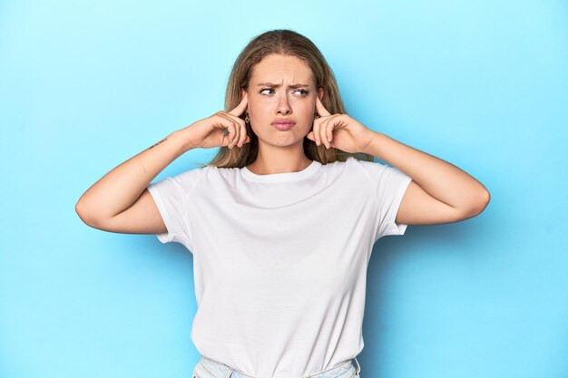 指で耳を覆う青い背景に白いTシャツを着た金髪の若い女性が強調された