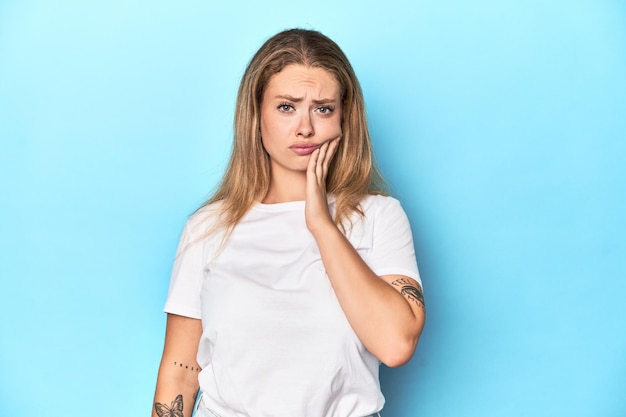 Foto giovane donna bionda in maglietta bianca su sfondo blu con un forte dolore ai denti mal di mola