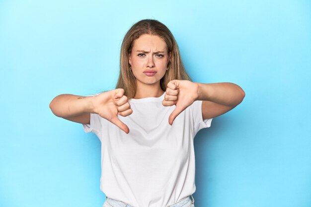 Photo blonde young woman in white tshirt on blue background showing thumb down and expressing dislike